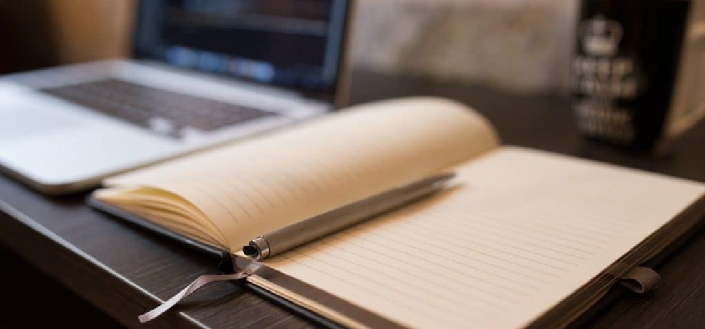 Open notebook with blank page, pen lying on top and laptop in background.