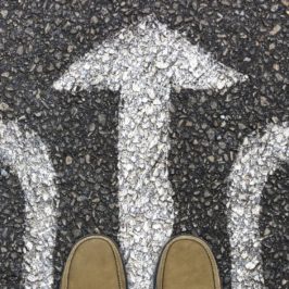 Toes of brown shoes on three white chalk arrows on road