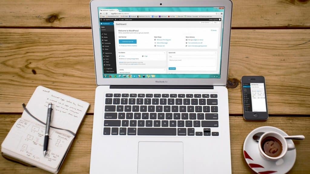 Open laptop with WordPress on screen, flanked by a notepad and pen, phone and cup of coffee