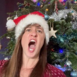 Debbie Emmitt in Santa hat, singing in front of Christmas tree