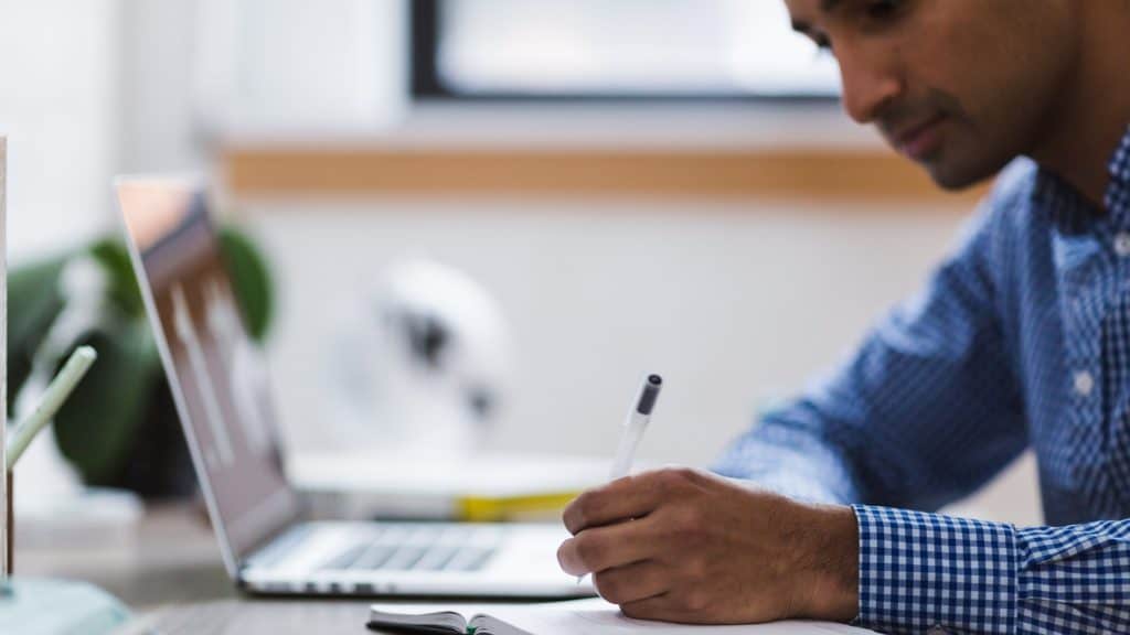 Male editor writing in notebook next to laptop