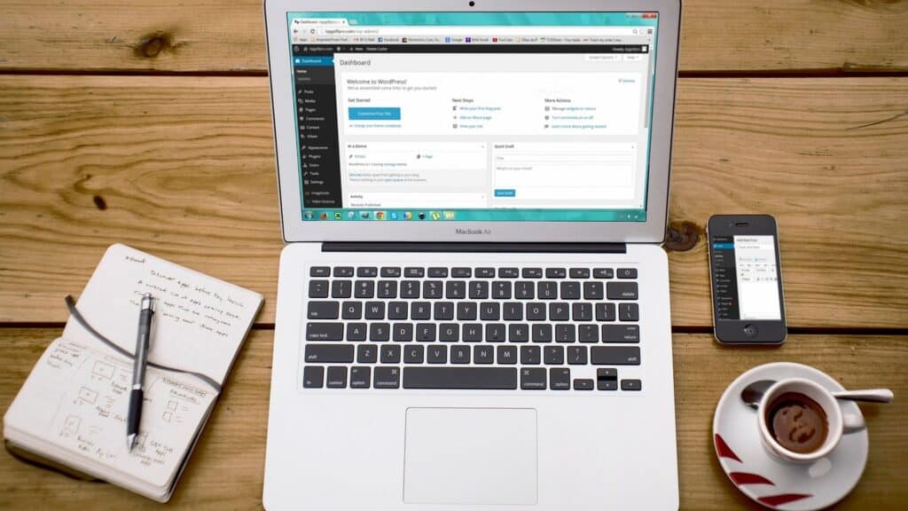 Open Macbook on wooden desk, WordPress on screen, with cup of coffee and notebook next to it