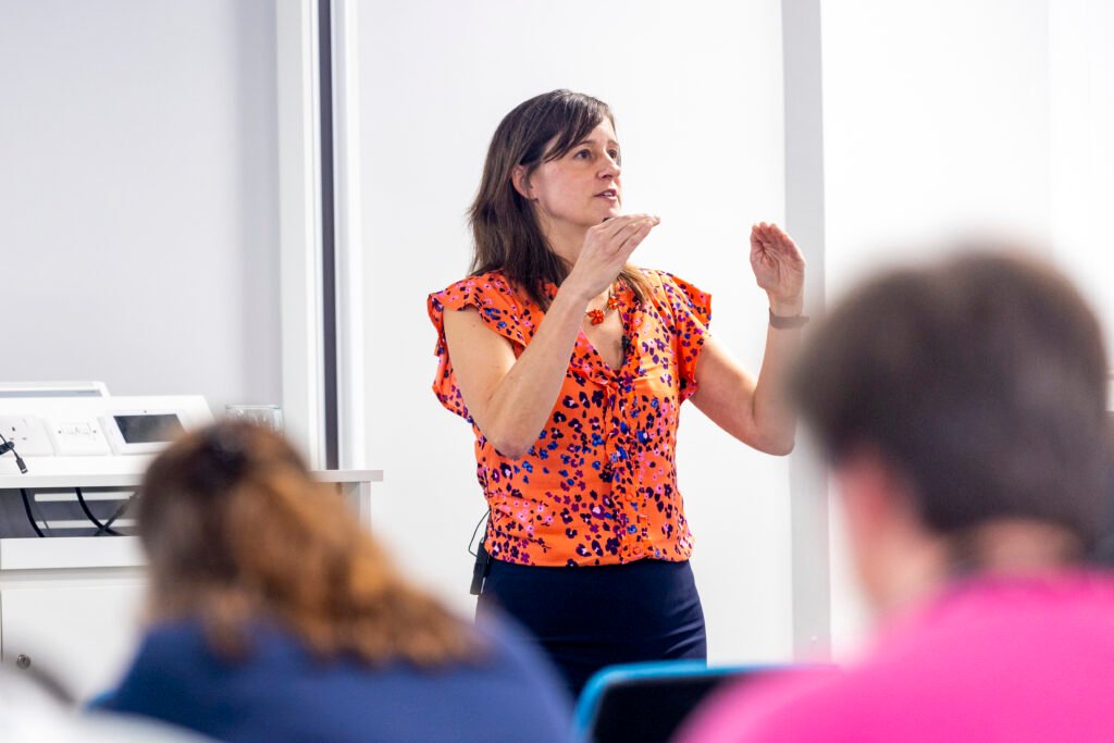 Woman with brown hair wearing orange blouse speaking in front of people