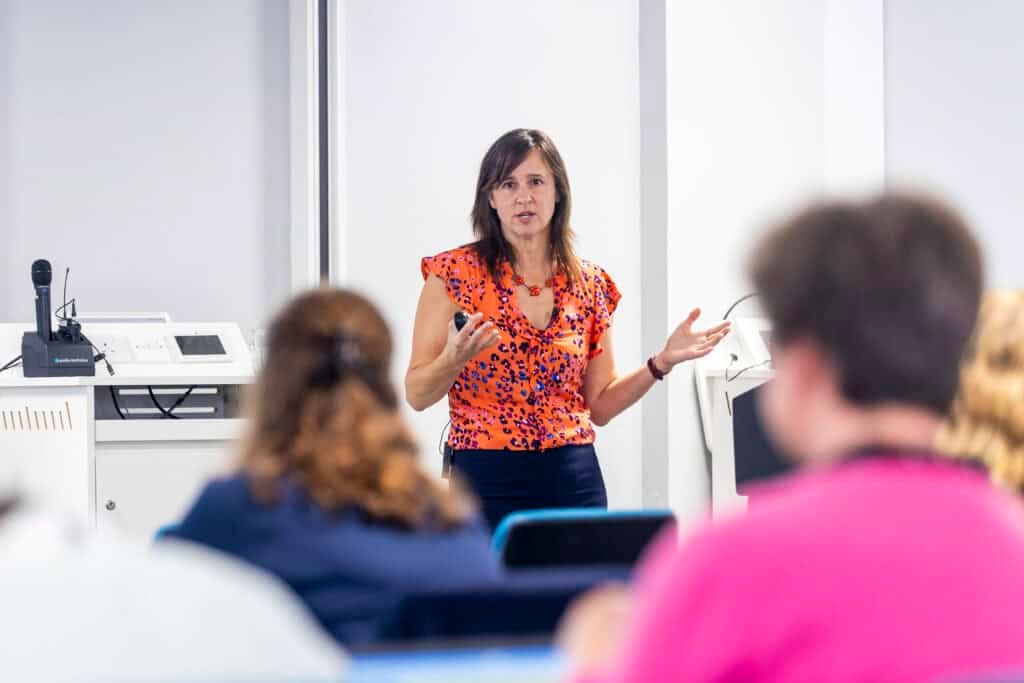 Woman with brown hair wearing orange blouse speaking in front of people