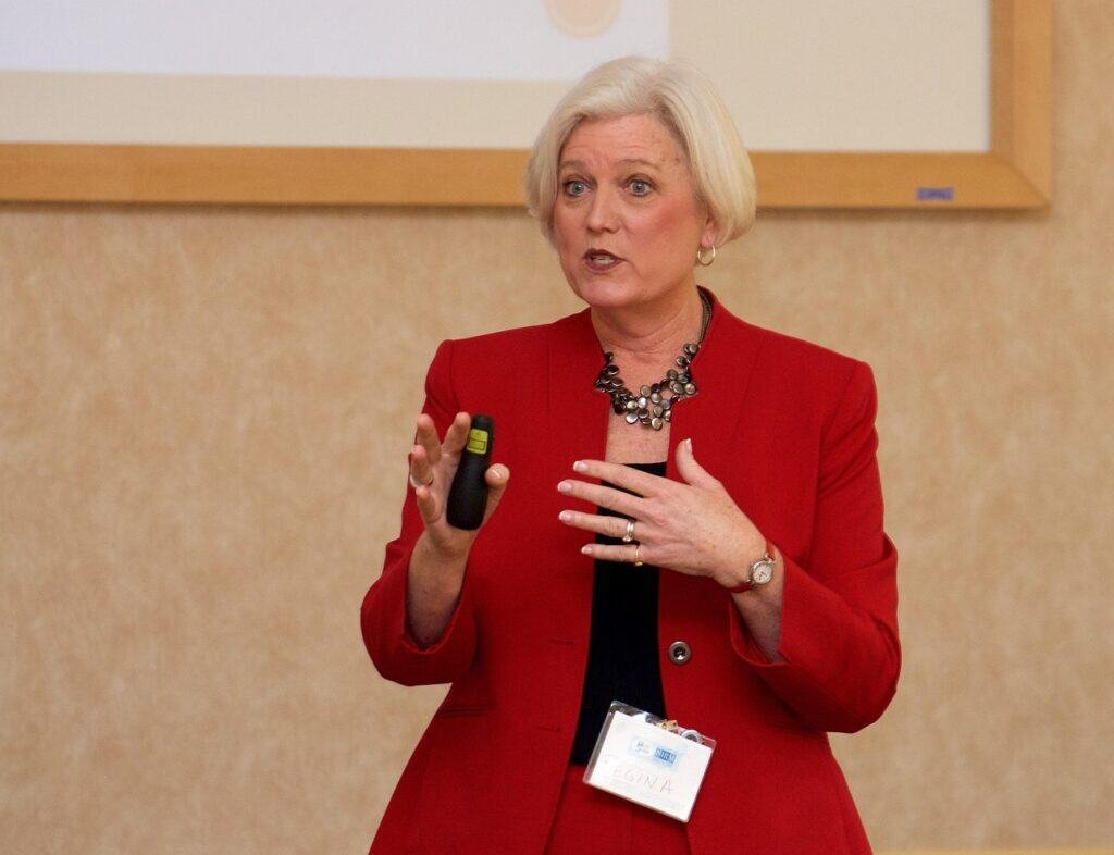 Woman with grey hair in smart red jacket speaking at the front of a room