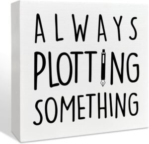 White square wooden desk sign with text "Always plotting something"